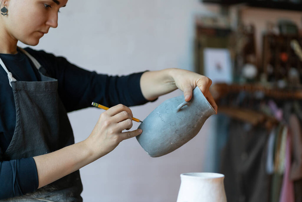Young serious skilled woman artisan with clay blue vase and pencil painting handmade earthenware while standing by workplace. Creative craft concept. Focused craftswoman makes sketches on ceramic jug. - Foto, Imagem