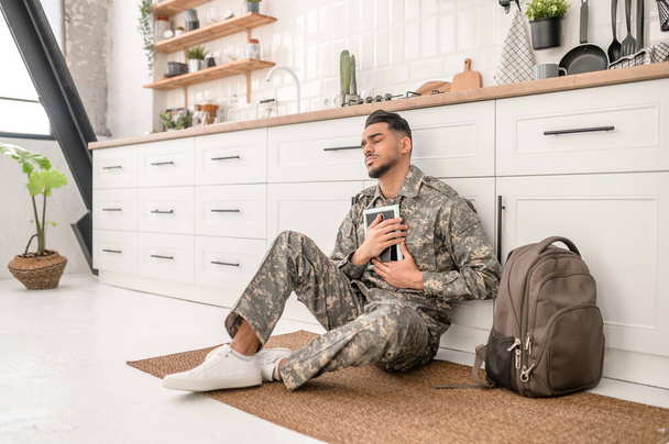 Sad military man seated on the kitchen floor pressing the framed photo against his chest - Photo, Image