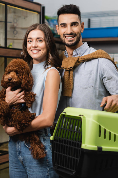 Cheerful multiethnic couple holding poodle and animal cage in pet shop  - Fotografie, Obrázek