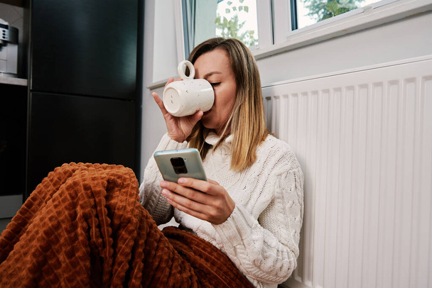 Worried sad woman sits under blanket near heating radiator and use smartphone, Rising costs in private households for gas bill due to inflation and war, Energy crisis - Foto, Bild
