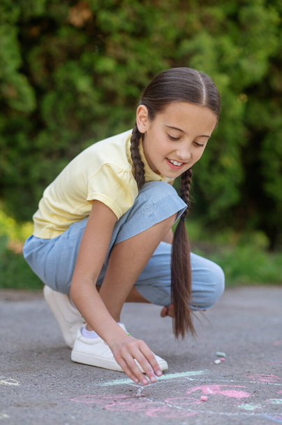 Lucky drawing. Dark-haired smiling girl with pigtails in tshirt and breeches carefully examining drawing on pavement - Zdjęcie, obraz