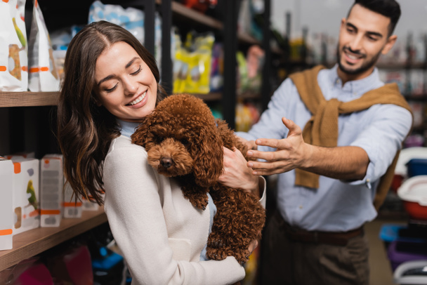 Smiling woman holding poodle near blurred arabian boyfriend and packages in pet shop  - Foto, afbeelding