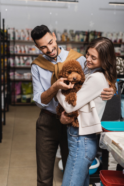 Cheerful multiethnic couple holding poodle in pet shop  - Photo, image