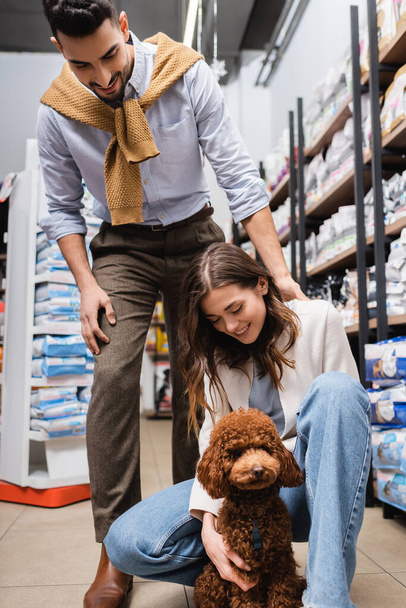 Cheerful interracial couple looking at brown poodle in pet shop  - Fotó, kép