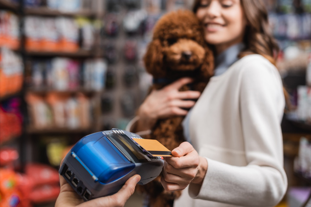 Размытая женщина держит пуделя и платит кредитной картой в зоомагазине  - Фото, изображение