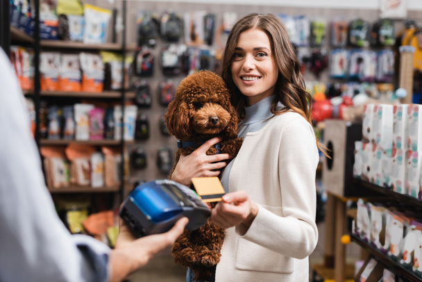 Smiling customer paying with credit card and holding poodle in pet shop  - Foto, Imagem