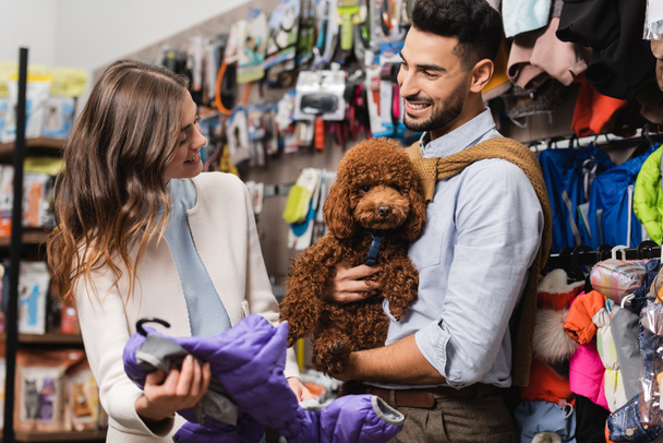 Smiling woman holding animal jacket near arabian boyfriend with poodle in pet shop - Valokuva, kuva