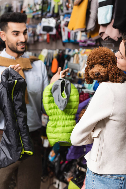 Woman holding brown poodle near blurred muslim boyfriend with animal jackets in pet shop  - Foto, afbeelding