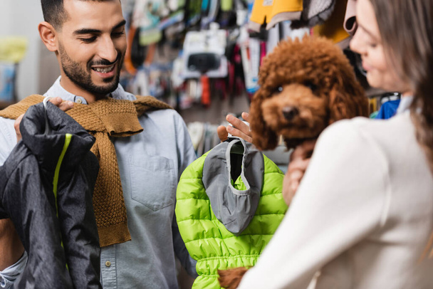 Smiling muslim man holding animal jackets near blurred girlfriend with poodle in pet shop - Foto, Imagem