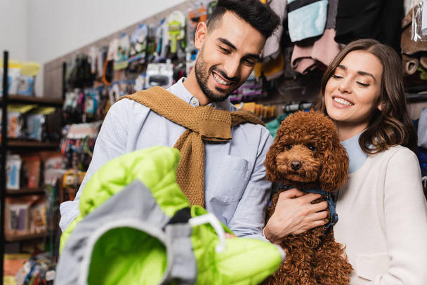 Cheerful multiethnic couple with poodle choosing animal jacket in pet shop  - Valokuva, kuva