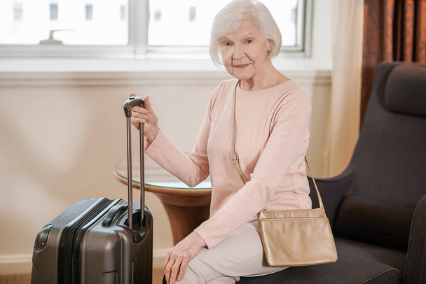 In a hotel. Good-looking senior woman sitting with a suitcase in a hotel room - Zdjęcie, obraz