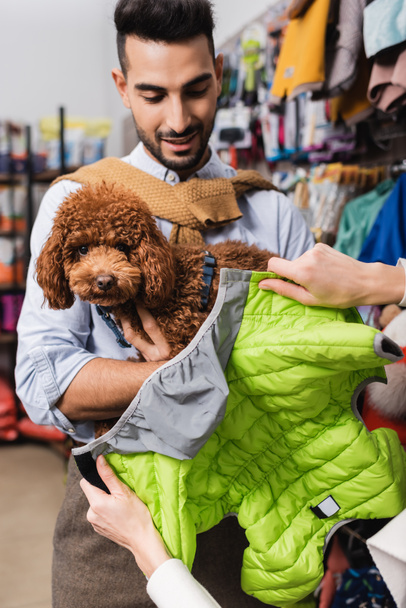 Woman holding animal jacket near arabian boyfriend with poodle in pet shop  - Foto, imagen