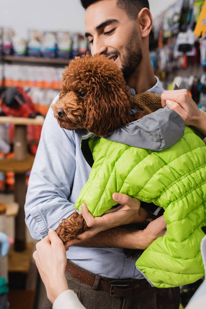 Woman wearing animal jacket on poodle near arabian boyfriend in pet shop  - Fotoğraf, Görsel