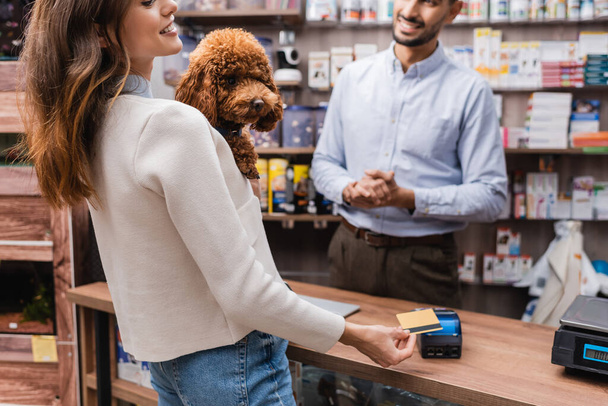 Cropped view of smiling woman holding poodle and paying with credit card near muslim seller in pet shop  - Fotografie, Obrázek