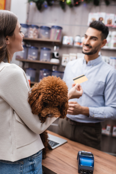 Smiling woman holding poodle and blurred blurred credit card near arabian seller in pet shop - Foto, afbeelding