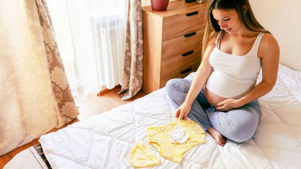 妊娠中のバッグベビー服。黄色の赤ん坊の服を保持する幸せな若い妊婦。マザーパッキングスーツケース、妊婦病院用バッグ。妊娠・産前・出産への期待 - 写真・画像