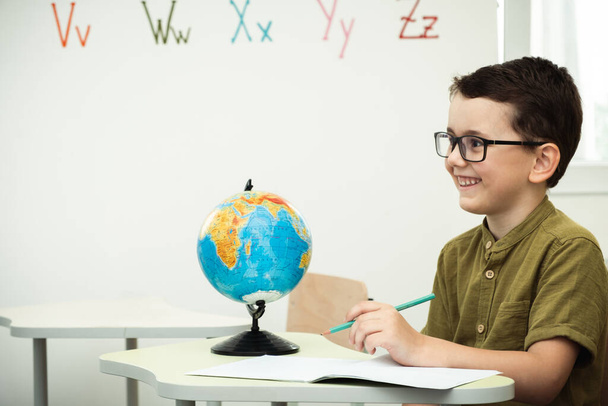 Cute caucasian schoolboy wearing glasses and sits at a desk in the classroom at elementary school. Little boy next the Globe at school. Back to school concept - Foto, Bild