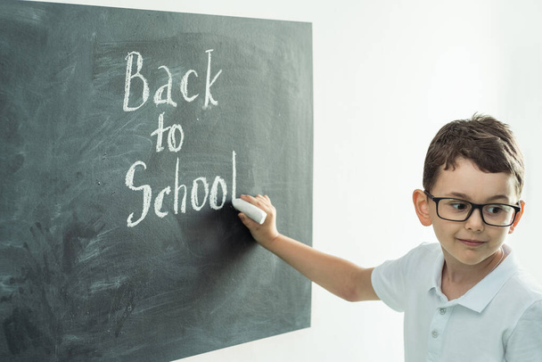 Χαριτωμένο καυκάσιο σχολιαρόπαιδο που φοράει γυαλιά και γράφει πίσω στο σχολείο κείμενο στο μαύρο σχολικό συμβούλιο στην τάξη στο δημοτικό σχολείο. - Φωτογραφία, εικόνα