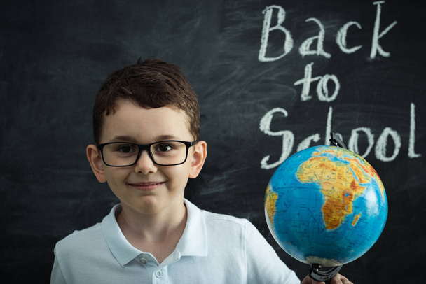 Cute caucasian schoolboy wearing glasses and holding the Globe against the black school board in the classroom at elementary school. Back to school concept - Fotografie, Obrázek