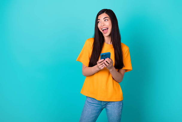 Photo of impressed brunette lady hold telephone look promo wear orange t-shirt isolated on turquoise color background. - Photo, Image