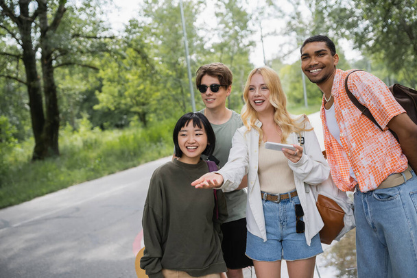smiling woman with smartphone pointing with hand near interracial friends on road - Photo, Image