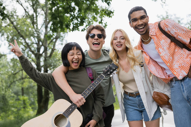 excited asian woman laughing near happy interracial friends with guitar and backpacks - Fotografie, Obrázek