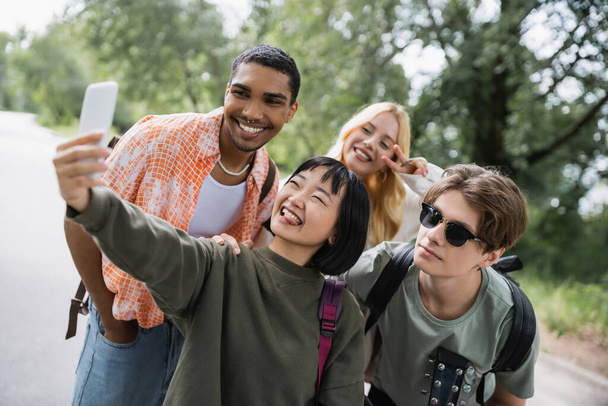 cheerful asian woman sticking out tongue while taking selfie with happy multiethnic friends - Valokuva, kuva