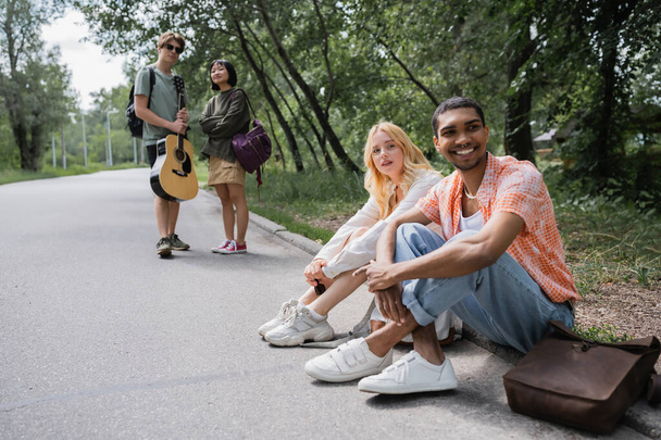 smiling interracial travelers sitting on road near blurred friends with guitar - Fotoğraf, Görsel