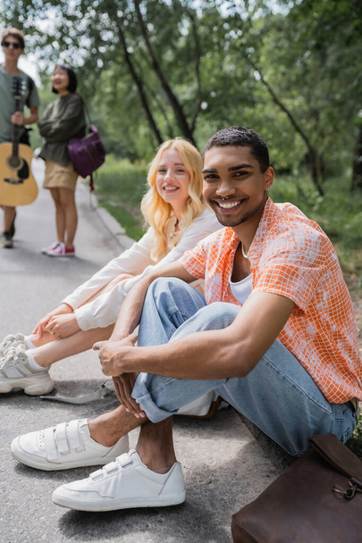 happy african american man smiling at camera while sitting on road near interracial friends - Foto, afbeelding