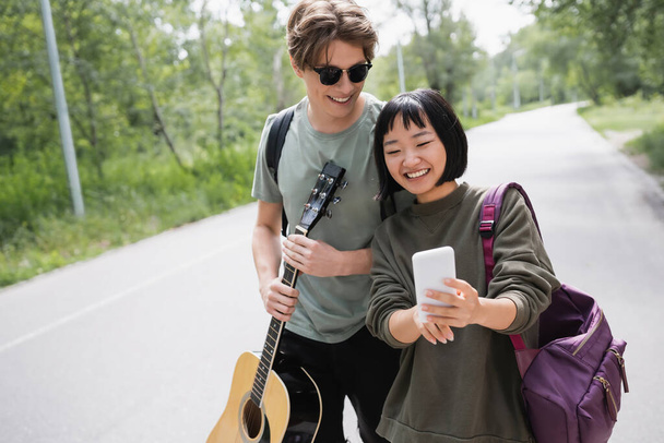 asian woman searching route on smartphone near boyfriend with acoustic guitar - Foto, Imagem