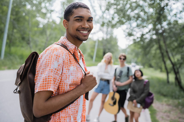 young african american man with backpack smiling at camera near friends on blurred background - Fotoğraf, Görsel