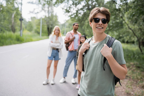 smiling man with sunglasses and backpack near multiethnic friends on blurred background - Photo, image