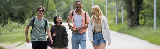 young and cheerful interracial friends walking on road near forest, banner - Photo, Image