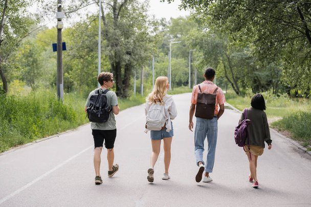 πίσω άποψη των νέων πολυεθνικών τουριστών περπάτημα στο δρόμο κοντά στο δάσος - Φωτογραφία, εικόνα