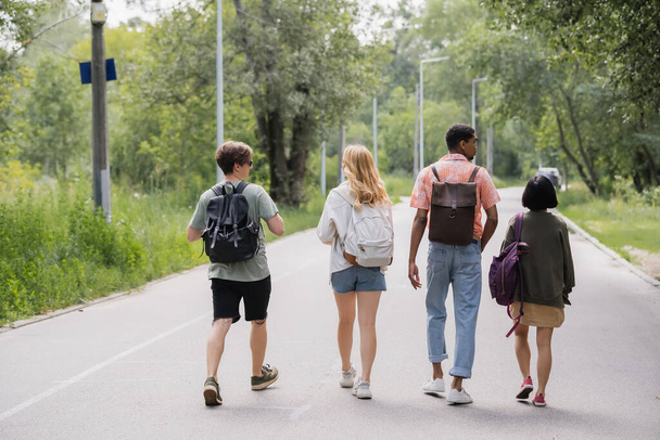вид ззаду на багатокультурних друзів з рюкзаками, що йдуть по сільській дорозі
 - Фото, зображення