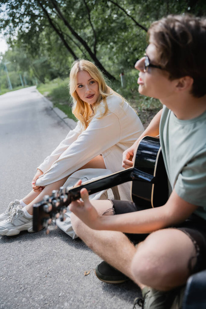 blurred man sitting on road near blonde woman and playing acoustic guitar - Photo, Image