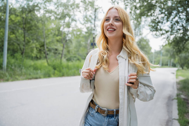 cheerful blonde woman with backpack looking away on countryside road - Foto, immagini