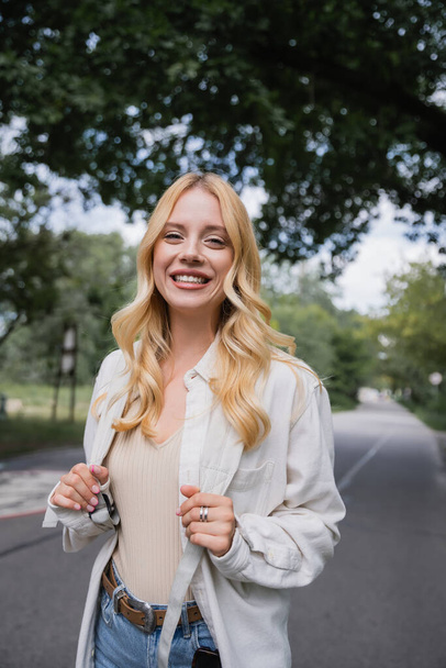 cheerful blonde traveler with backpack smiling at camera on blurred road - Foto, afbeelding