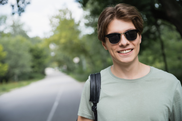 улыбающийся турист в солнечных очках, смотрящий в камеру на размытой дороге - Фото, изображение
