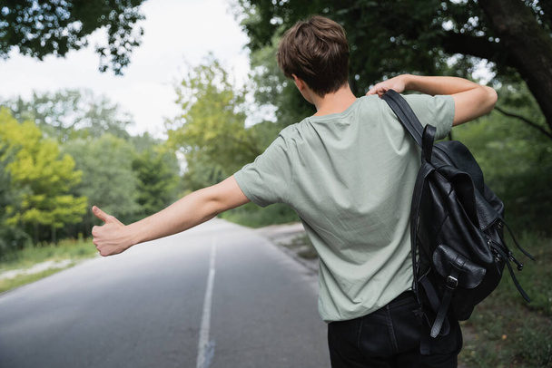 back view of hitchhiker with backpack stopping car with thumb up on blurred road - Foto, Imagem