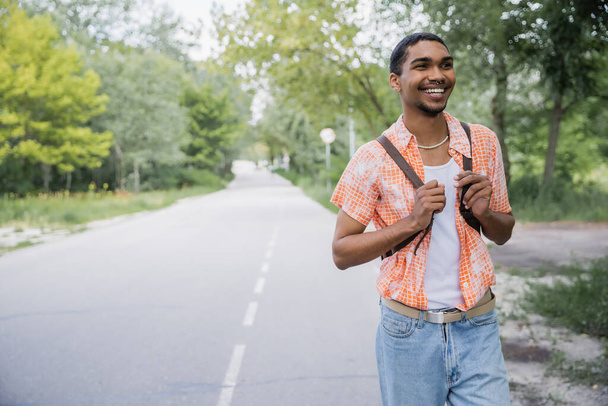 щасливий афроамериканський мандрівник з рюкзаком, що йде по дорозі біля лісу
 - Фото, зображення