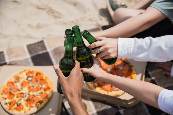 partial view of multiethnic friends clinking beer bottles near tasty pizza on beach - 写真・画像