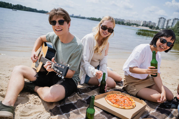 щасливий чоловік грає на гітарі біля щасливих міжрасових жінок під час пляжної вечірки
 - Фото, зображення