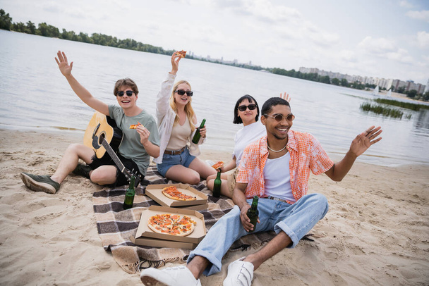 cheerful multiethnic friends waving hands during picnic on riverside - Fotó, kép