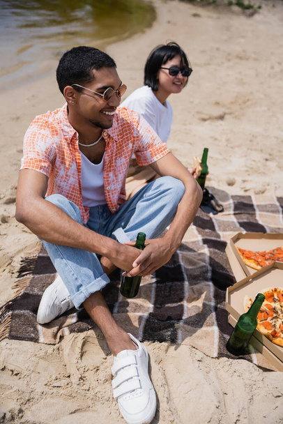 smiling interracial friends sitting with beer and pizza on blanket near river - Foto, afbeelding