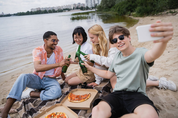 smiling man taking selfie with multiethnic friends clinking beer bottles near pizza - Photo, Image
