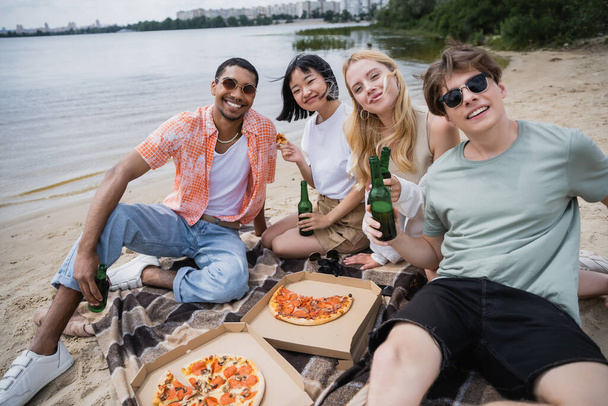 νέους πολυεθνικούς φίλους κοιτάζοντας κάμερα κατά τη διάρκεια πάρτι στην παραλία με μπύρα και πίτσα - Φωτογραφία, εικόνα