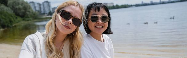 cheerful interracial women in sunglasses looking at camera near river, banner - 写真・画像