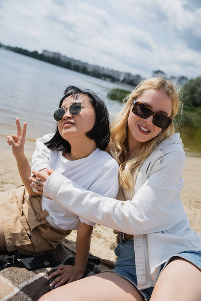 blonde woman in sunglasses embracing asian friend showing victory sign on beach - Foto, afbeelding