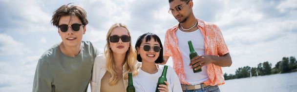 happy multiethnic friends holding beer near river on summer day, banner - 写真・画像